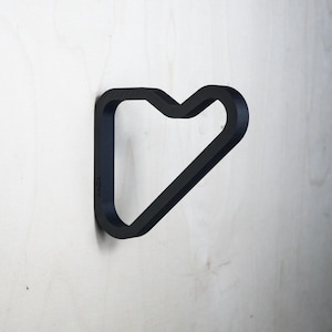 May include: A black, heart-shaped wall hook mounted on a light-colored wooden surface. The hook is made of a thick, solid material and has a minimalist design. The hook is a decorative item that can be used to hang items.