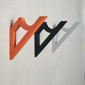 May include: Three wall-mounted coat hooks in orange, black, and gray. Each hook has a triangular shape with a curved top section. The orange hook has the text "HAGA Design" printed on it. The hooks are mounted on a light-colored wooden surface.