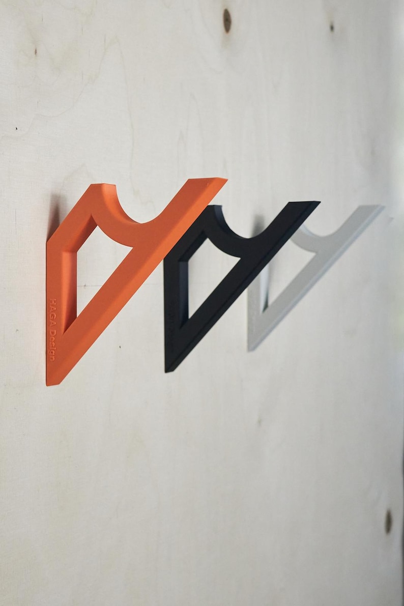 May include: Three wall-mounted coat hooks in orange, black, and gray. Each hook has a triangular shape with a curved top section. The orange hook has the text "HAGA Design" printed on it. The hooks are mounted on a light-colored wooden surface.