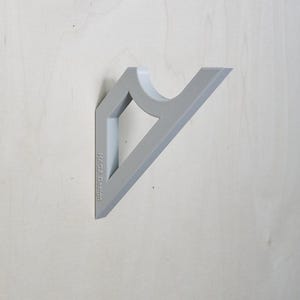 May include: A gray wall-mounted shelf bracket with a geometric design. The bracket has a triangular shape with a curved section and a rectangular opening. The text "HAGA Design" is printed on the side.