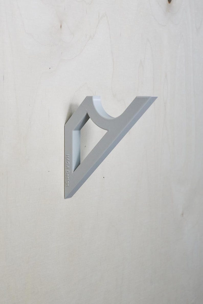 May include: A gray wall-mounted shelf bracket with a geometric design. The bracket has a triangular shape with a curved section and a rectangular opening. The text "HAGA Design" is printed on the side.