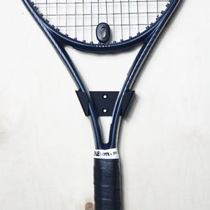 May include: A navy blue tennis racket with a black grip and a black wall mount. The racket has a white label with the text "Wilson Pro". The strings are silver.
