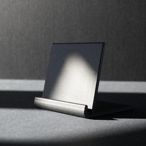 May include: A minimalist, dark gray metal card holder. The holder has a rectangular back and a curved base, designed to hold a card upright. The image is lit with a spotlight, creating shadows.