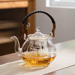 May include: A clear glass teapot with a curved spout and a wooden handle wrapped in black material. The teapot contains a golden-colored liquid and has a glass infuser inside. Two small fish-shaped charms hang from the handle.