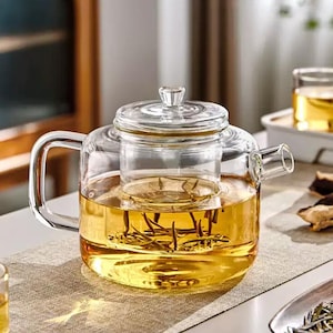 May include: A clear glass teapot with a built-in infuser, filled with golden tea. The teapot has a handle and spout, and a clear lid. The tea infuser is visible inside the teapot. The teapot is sitting on a neutral-colored surface.