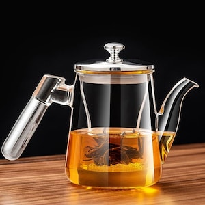 May include: A clear glass teapot with a stainless steel lid and handle. The teapot is filled with amber-colored tea and tea leaves. The teapot is sitting on a wooden surface.