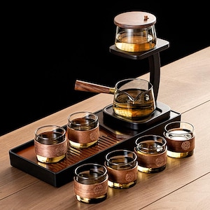 Può includere: Un moderno set da tè con una teiera in vetro con manico in legno, un infusore in vetro con coperchio in legno e sei bicchieri piccoli con dettagli in rame e oro. Il set è disposto su un vassoio e un supporto in legno.
