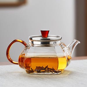 Op de afbeelding: Een heldere glazen theepot met een geometrisch ontwerp, gevuld met amberkleurige thee en theebladeren. Het heeft een zilveren deksel met een rode knop en een schildpadkleurige handgreep. De theepot staat op een licht oppervlak.