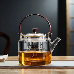 May include: A clear glass teapot with a brown wooden handle and lid. The teapot has a built-in infuser and is filled with amber-colored liquid. The teapot is sitting on a light-colored surface.