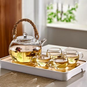 May include: A clear glass tea set with a woven handle on the teapot. The set includes a teapot filled with tea and four small teacups, all resting on a white and wood-tone tray. The tea is a light amber color.