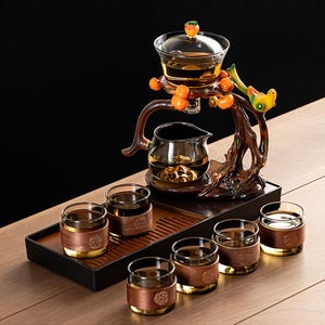 May include: A glass tea set with a decorative wooden stand featuring a bird and fruit design. The set includes a teapot, a serving pot, six small glasses, and a tray. The tea set is designed for serving tea.