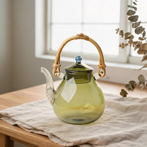 Peut inclure: Une théière en verre vert olive translucide dotée d'un bec verseur incurvé avec des détails nervurés, d'un couvercle assorti avec un petit bouton sphérique bleu et d'une anse en rotin arrondie fixée par des boucles tressées.