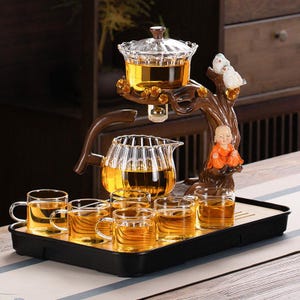 Puede incluir: Un juego de té con una tetera de vidrio, tazas y una bandeja de servicio. El juego incluye una tetera de vidrio, una taza de té de vidrio y un soporte de madera decorativo con una pequeña estatua de un monje y dos pájaros. El té es de color ámbar.