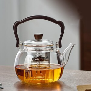May include: A clear glass teapot with a dark brown handle and lid. The teapot is filled with amber-coloured tea, and has a built-in infuser. The teapot is designed for brewing loose leaf tea.