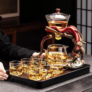 Può includere: Un set da tè con una figura decorativa a forma di scimmia. Il set include una teiera in vetro, una brocca e diverse tazzine da tè, tutte piene di tè. Il set è presentato su un vassoio nero.