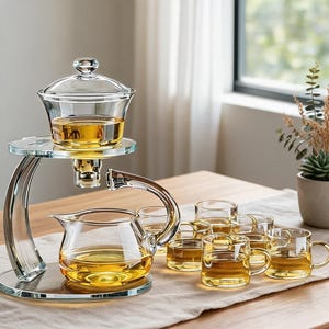 Könnte beinhalten: Ein Teeservice aus klarem Glas mit einem Etagenständer, einem oberen Brühgefäß und einer unteren Kanne. Das Set enthält sechs kleine Glastassen mit Henkeln, die mit bernsteinfarbenem Tee gefüllt und auf einer ebenen Fläche angeordnet sind.
