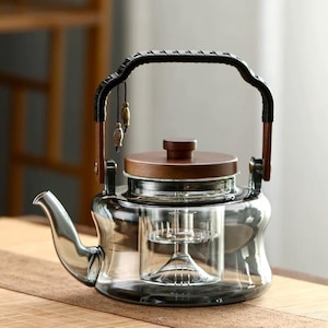 May include: A clear glass teapot with a wooden lid and handle. The handle is wrapped in black material. The teapot has a built-in infuser and decorative fish charms hanging from the handle. The teapot is designed for brewing tea.