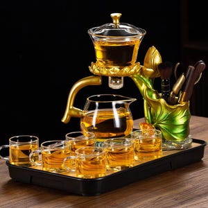 May include: A tea set with a golden and glass design. The set includes a tea pot, tea cups, and a tea infuser. The tea set is on a black tray. The tea set has a gold lotus flower design.