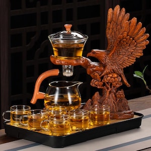 Puede incluir: Un juego de té de madera con un águila tallada, una tetera de vidrio y varias tazas de té pequeñas llenas de té. El juego incluye una bandeja y un infusor de té de vidrio. El águila está posada en una rama.