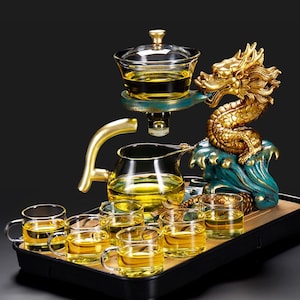 Può includere: Servizio da tè con drago dorato, composto da teiera, tazze e vassoio. Il drago è appoggiato su una base di onde turchesi e dorate. Il set include un infusore in vetro e un vassoio con sei piccole tazze di vetro. Il tè è di colore giallo dorato.