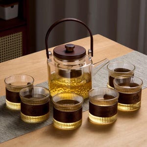 May include: A glass teapot with a wooden handle and lid, filled with tea, is surrounded by six matching glasses. Each glass has a dark brown band and a ribbed design, creating a warm, inviting tea set.