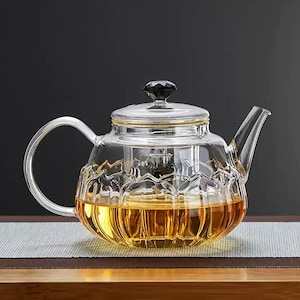 Puede incluir: Una tetera de vidrio transparente con asa y boquilla curvadas. La tetera tiene un diseño decorativo y texturizado en la mitad inferior y un pomo negro en forma de gema en la tapa. La tetera está llena de un líquido dorado.