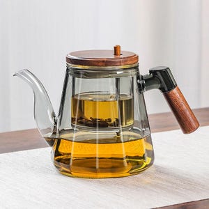 May include: A clear glass teapot with a wooden lid and handle. The teapot has a built-in infuser and is filled with tea. The handle is made of wood and black plastic. The teapot is sitting on a white surface.