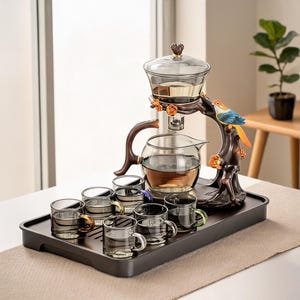 May include: A tea set featuring a glass teapot and six small glass cups on a dark tray. The teapot sits on a brown, tree-shaped stand decorated with orange flowers and a colorful bird figurine. The cups have small, differently colored handles.