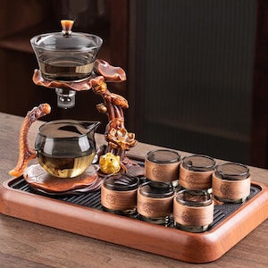 Pode incluir: Um conjunto de chá completo com uma bandeja de madeira escura. O conjunto inclui uma chaleira de vidro, um recipiente para servir e seis xícaras pequenas. A chaleira e as xícaras têm um tom escuro. O conjunto é suportado por um suporte decorativo de madeira com detalhes florais e de peixe.