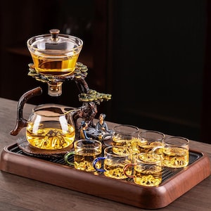 Op de afbeelding: Een glazen theeset met een decoratieve boomvormige standaard. De set bevat een theepot, een serveervaas en verschillende kleine kopjes, allemaal gevuld met thee. De set wordt gepresenteerd op een houten dienblad, met miniatuurbeeldjes.