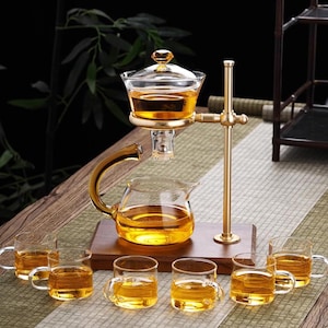 Puede incluir: Un juego de té de vidrio con un soporte de metal dorado y una base de madera. El juego incluye una tetera, una cámara de elaboración con tapa y seis tazas de té pequeñas, todas llenas de té de color ámbar. El juego está diseñado para preparar y servir té.