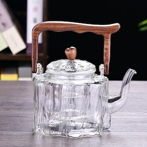 Peut inclure: Une théière en verre transparent avec une poignée et un couvercle en bois. La théière présente un design texturé unique et un bouton en bois en forme de cœur sur le couvercle. Le bec verseur est incurvé.
