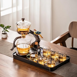 May include: A glass tea set featuring a clear teapot with a lid, a glass pitcher, and six small glass cups. The set is supported by a dark brown branch-shaped stand adorned with a colorful bird figurine and orange flower accents on a tray.