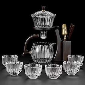 May include: A clear glass tea set featuring a teapot, infuser, and six small cups. The teapot and cups have a ribbed design. The set includes a wooden stand and serving tools, set against a dark background.