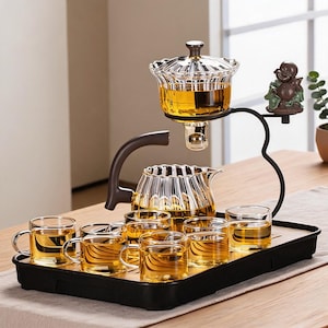 Puede incluir: Un juego de té de cristal que incluye una tetera estriada, un infusor de cristal suspendido con tapa y seis tazas pequeñas de cristal. El conjunto está dispuesto sobre una bandeja rectangular negra con una base de madera y una figura.