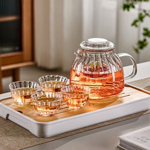 May include: A clear glass tea set with a teapot and four small cups, all filled with a reddish-brown liquid. The teapot has a lid and a handle. The set sits on a bamboo tray with a white border.