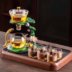 Puede incluir: Un juego de té con una tetera de vidrio y seis tazas pequeñas, todas llenas de té. El juego incluye un soporte decorativo con un diseño de flor de loto y una bandeja de madera. La tetera tiene un asa dorada y las tazas tienen fundas de madera.