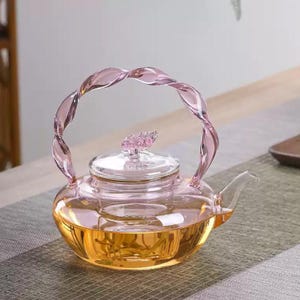 Puede incluir: Una tetera de vidrio transparente con un asa rosa retorcida y un diseño floral rosa en la tapa. La tetera está llena de un líquido dorado, probablemente té. La tetera está sobre una superficie gris y verde.