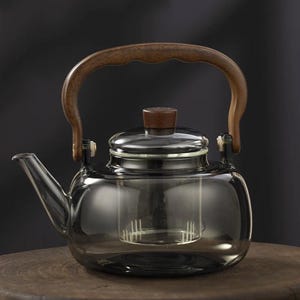 Op de afbeelding: Een heldere glazen theepot met een houten handvat en deksel. De theepot heeft een ingebouwde infuser en staat op een houten oppervlak. De handgreep en de knop van het deksel zijn van donker hout. De theepot is ontworpen voor het zetten van thee.