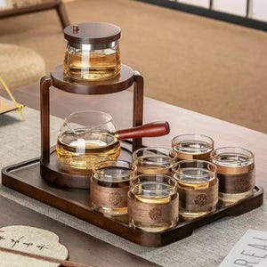 Puede incluir: Un juego de té de madera con una tetera de vidrio, un servidor de vidrio y seis vasos pequeños. El juego de té está sobre una bandeja de madera. La tetera y el servidor tienen asas de madera. Los vasos tienen detalles de madera. El té es de color ámbar.
