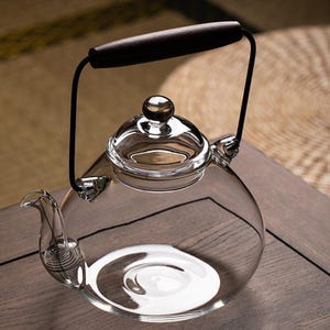 May include: A clear glass teapot with a dark wooden handle and a black metal frame. The teapot has a rounded body, a spout, and a lid with a small knob. The teapot is sitting on a wooden surface.