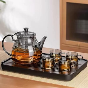 Może przedstawiać: Szklany zestaw do herbaty z czajniczkiem z wbudowanym zaparzaczem i czterema pasującymi filiżankami, wszystkie wypełnione herbatą. Czajniczek i filiżanki znajdują się na czarnej tacy, do serwowania herbaty.