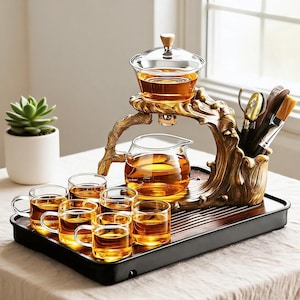 Puede incluir: Un juego de té con una tetera de cristal y seis tazas pequeñas de cristal llenas de té ámbar. El conjunto incluye un soporte de color bronce con forma de rama de árbol, una bandeja rectangular oscura y un soporte para utensilios de té.