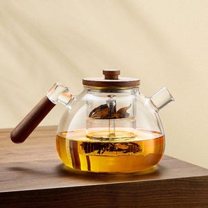 May include: A clear glass teapot with a wooden handle and lid. The teapot is filled with a golden-colored liquid and tea leaves. The teapot sits on a wooden surface.
