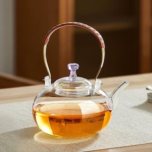 May include: A clear glass teapot with a brown leather-wrapped handle and a purple lid. The teapot is filled with amber-colored liquid, likely tea. The design is elegant and suitable for serving tea.