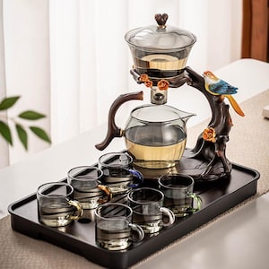 Pode incluir: Um conjunto de chá com uma chaleira de vidro, um infusor e seis chávenas pequenas. A chaleira e o infusor estão sobre um suporte de madeira decorativo com uma figura de pássaro. As chávenas têm pegas coloridas e o conjunto está numa bandeja preta.