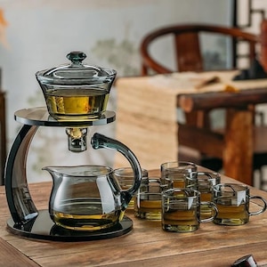 May include: A clear glass tea set, including a teapot, serving bowl, and six small teacups. The tea set is on a black stand. The tea is amber-coloured. The background features a wooden table and a blurred wooden chair.