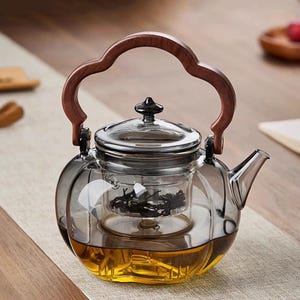 May include: A clear glass teapot with a wooden handle and lid. The teapot has a built-in infuser and is filled with tea. The handle is a dark brown color, and the lid is silver. The teapot is sitting on a light-colored surface.