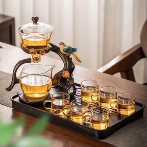 May include: A tea set with a glass teapot, a pitcher, and six small cups, all filled with tea. The set is on a black tray, with a decorative bird perched on a branch. The set is made of glass and dark brown wood.