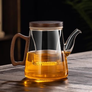 Könnte beinhalten: Eine Teekanne aus klarem Glas mit Holzgriff, Deckel und Sieb. Die Teekanne ist mit einer goldfarbenen Flüssigkeit gefüllt, wahrscheinlich Tee. Das Design ist modern und minimalistisch.